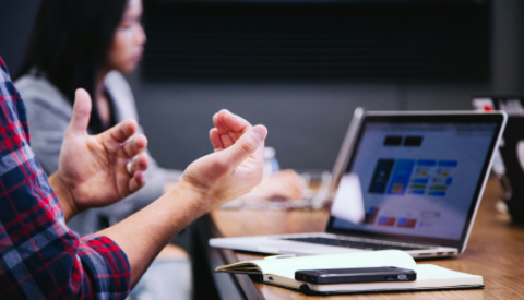 man gesturing at a desk with a laptop with woman in background